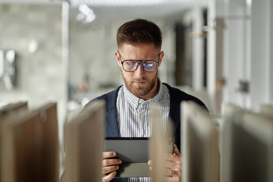 Caucasian young adult man wearing glasses using digital tablet, focusing on screen, short beard visible, modern office or showroom environment