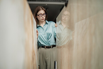Caucasian young adult woman wearing glasses examining large marble slabs in showroom, standing between stone panels, focused expression, hands touching surfaces, indoor setting