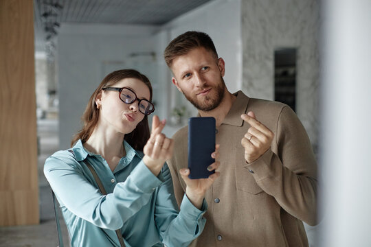 Caucasian young woman wearing glasses and Caucasian young man making finger heart gestures while taking selfie with smartphone, both looking at camera and smiling playfully - Powered by Adobe