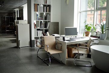 Modern office workspace featuring empty chairs surrounding desk with computer monitor, camera lens, potted plants and large windows overlooking urban street with brick building