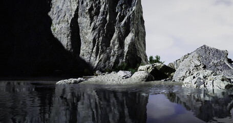 A large rocky mountain stands tall beside a calm lake, with gentle ripples creating soft reflections on the waters surface. Green plants peek through the rocks under a gray sky.
