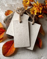 Two blank tags, tied with twine, rest on a stone, surrounded by autumn leaves and flowers