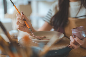 Hands of woman artist painting handmade ceramic product in pottery studio – creative process of decorating sculpture with brush for small business, art workshop, and modern craft design startup