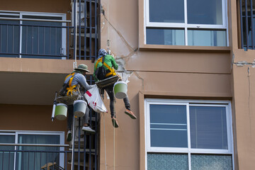 Workers repairing building facade with safety harnesses, using buckets and tools. Urban maintenance scene