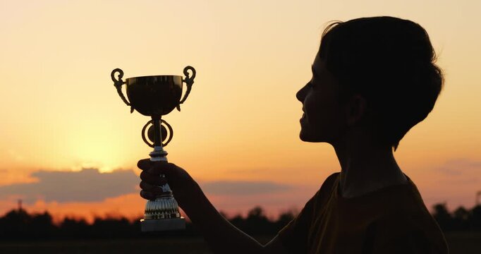 Child holding trophy celebrates victory on sunset background. Child joyfully lifts trophy with pride. Young champion shines against evening sky. Child success achievement. Winning kid.