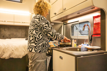 Woman preparing food in a cozy campervan kitchen with bed in the background during a motorhome road...