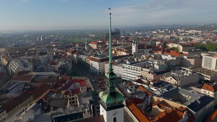 High aerial drone flyby, closely skimming the historic spire of St. Jacob's Church, overlooking the central Brno cityscape in South Moravia, Czechia, on a sunny spring afternoon.