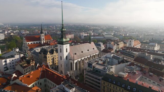 Aerial drone circling shot of the historic St. Jacob's Church spire soaring above the Brno cityscape in the South Moravian region of Czechia on a bright spring day.