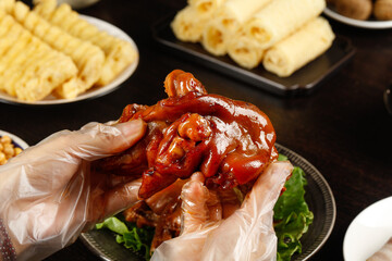 Glazed Pork Trotters Held in Gloved Hand with Traditional Asian Snacks and Side Dishes