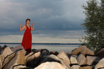 Pretty young brunette woman in red dress walking by rocks at the seashore