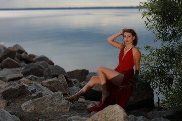Slender long legged barefoot beautiful brunette young woman in long red dress sitting on the rocks of the sea coastline