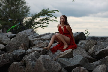 Slender long legged barefoot beautiful brunette young woman in long red dress sitting on the rocks of the sea coastline