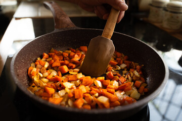 Close-up of woman's hand frying onions and carrots in a hot frying pan with vegetable oil on the induction stove, homemade cooking concept