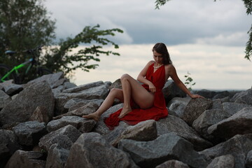 Slender long legged barefoot beautiful brunette young woman in long red dress sitting on the rocks of the sea coastline