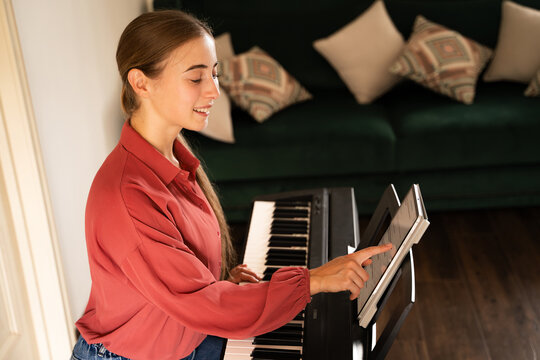 Side view portrait of smiling young woman with tablet taking online piano lessons at home, distance learning of new hobbies with digital classical piano indoors in daylight