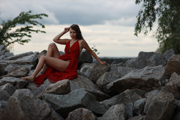 Slender long legged barefoot beautiful brunette young woman in long red dress sitting on the rocks of the sea coastline