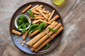 Plate with various breadsticks, basil oil and fresh basil on a brown granite background, horizontal shot, above view