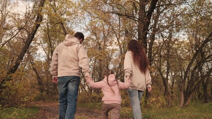 mom dad and child are walking along forest autumn road, happy family life, yellow dry leaves, fallen leaves in the outdoor nature park, father mother and kid are walking, girl with parents together