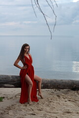 Young long-legged woman in a long red dress sitting on a wooden block on a sandy seashore
