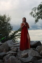 Naklejka premium Pretty slim brunette woman in red dress standing full length on the rocks near green bushes