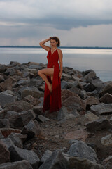 Beautiful young brunette woman in long red dress standing on the rocks at the sea