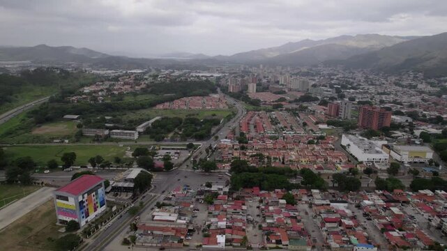 sweeping drone shot capturing Maracay, Venezuela, where modern buildings and dense housing transition into lush green foothills. Contrast development and poverty