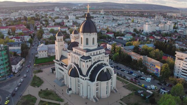 A cinematic aerial shot slowly pans right, showing a majestic Orthodox cathedral and the city of Targu Mures, Romania, bathed in the warm, glowing light of a golden hour sunset.
