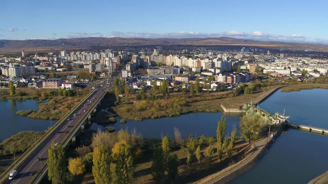 A cinematic pull-back aerial shot establishing the cityscape of Bacau, Romania. The view reveals traffic on a bridge and a dam on the river during a sunny autumn day.