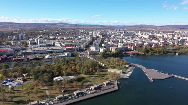 A slow-panning aerial drone shot reveals a sprawling cityscape on the shore of a large blue lake. Bacau. Autumn trees dot the waterfront park below, with mountains on the horizon.