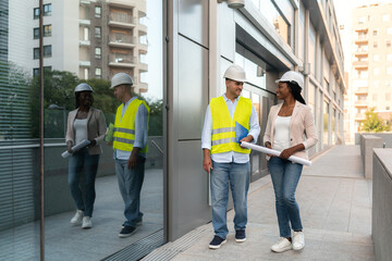 Two diverse Construction workers in safety helmets and vests walking and talking outside office