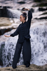 Fototapeta premium Woman practicing Tai Chi in nature, wearing a traditional black martial arts uniform, standing gracefully near a waterfall, focused on meditation and balance. Healthy lifestyle in asian culture