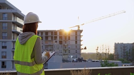 Construction female engineer taking notes and writing on clipboard while inspecting a building site at sunset. Industry development concept