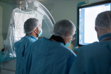 Medical team in scrubs and masks confer near a monitor in a hospital operating room. The team reviews data, preparing for or assessing the ongoing medical procedure