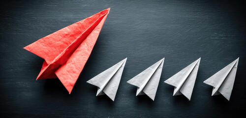 A red paper airplane leads a line of white paper airplanes on a dark surface