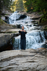 Beautiful young woman performing slow tai chi movements near a waterfall and river at sunset capturing harmony balance and fitness in nature.