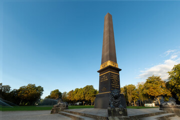 A tall obelisk with golden decoration rises into the sky. At its base, lion statues flank the...