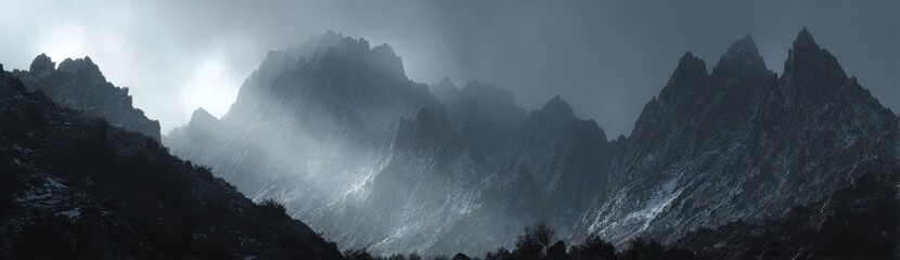 Misty mountain range with jagged peaks and light breaking through clouds, evoking a sense of awe and solitude in a grayscale scene