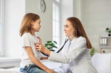 Fototapeta premium Female friendly doctor with stethoscope examining child's lungs, breathing and heart rate sitting on the couch at the hospital. Pediatrician woman checking a little patient boy in exam room.