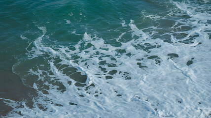 Obraz premium An aerial view of turquoise ocean waves creating white seafoam as they wash ashore on a sandy beach.