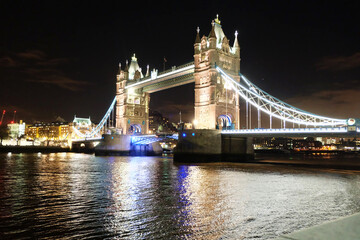 Tower Bridge in London illuminated at night with bright lights reflecting on the River Thames and a dark city skyline in the background