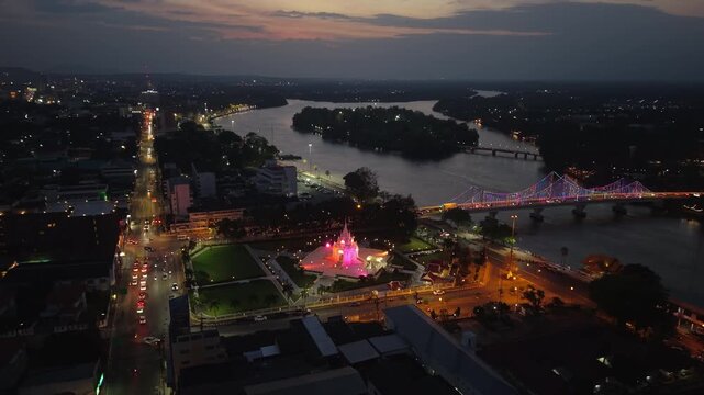 Night aerial of Surat Thani City Pillar Shrine glowing, with city lights, Tapi River, and traffic illuminating the urban landscape of Thailand.