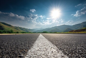 Long asphalt road stretches into the distance, toward rolling green hills under a bright sun in a partly cloudy blue sky