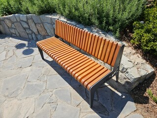A modern wooden and metal bench sits in a sunny park on a stone patio. Ideal for urban planning, landscape architecture, or content about city recreation and public spaces.