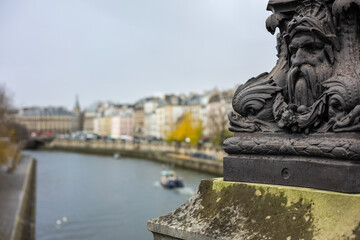 Close-up of an ornate, historic lamppost on the Pont Neuf bridge in Paris, France. The cast iron base features grotesque mascarons and sea creatures.
