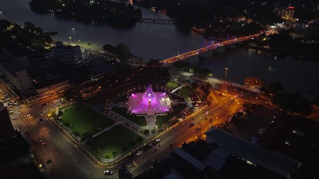 Night aerial of Surat Thani City Pillar Shrine glowing, with city lights, Tapi River, and traffic illuminating the urban landscape of Thailand.