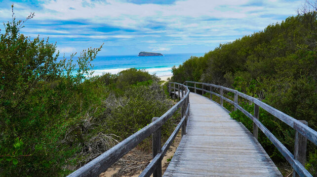 Scenic coastal walkway leading to a serene beach with distant island view under a cloudy sky.Access to the ocean Australia.