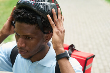 Young Black man adjusting bicycle helmet, wearing smartwatch and carrying insulated food delivery backpack, preparing for delivery outdoors on paved path in urban park setting