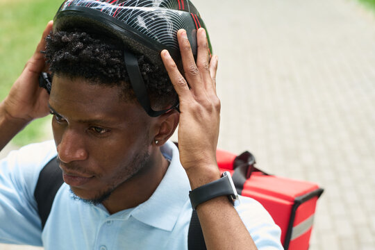 Young Black man adjusting bicycle helmet, wearing smartwatch and carrying insulated food delivery backpack, preparing for delivery outdoors on paved path in urban park setting