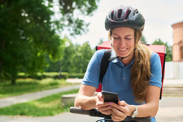 young woman wearing helmet smiling while using smartphone, delivering food on bicycle outdoors, carrying insulated backpack, looking down at device, urban park background