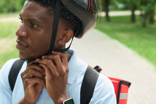 Young Black man fastening bicycle helmet strap while wearing delivery backpack outdoors, looking away from camera, preparing for work, smartwatch visible on wrist